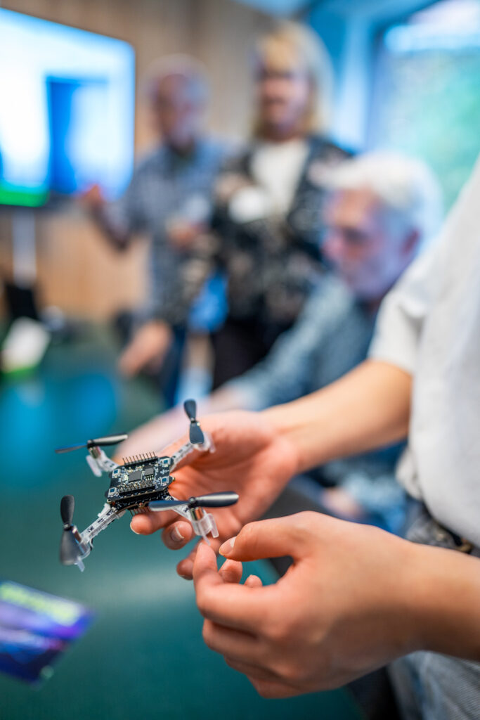 A person holds a small quadcopter drone, focussing on its details. In the background, three people are blurred, suggesting a group discussion or presentation in a modern indoor setting.