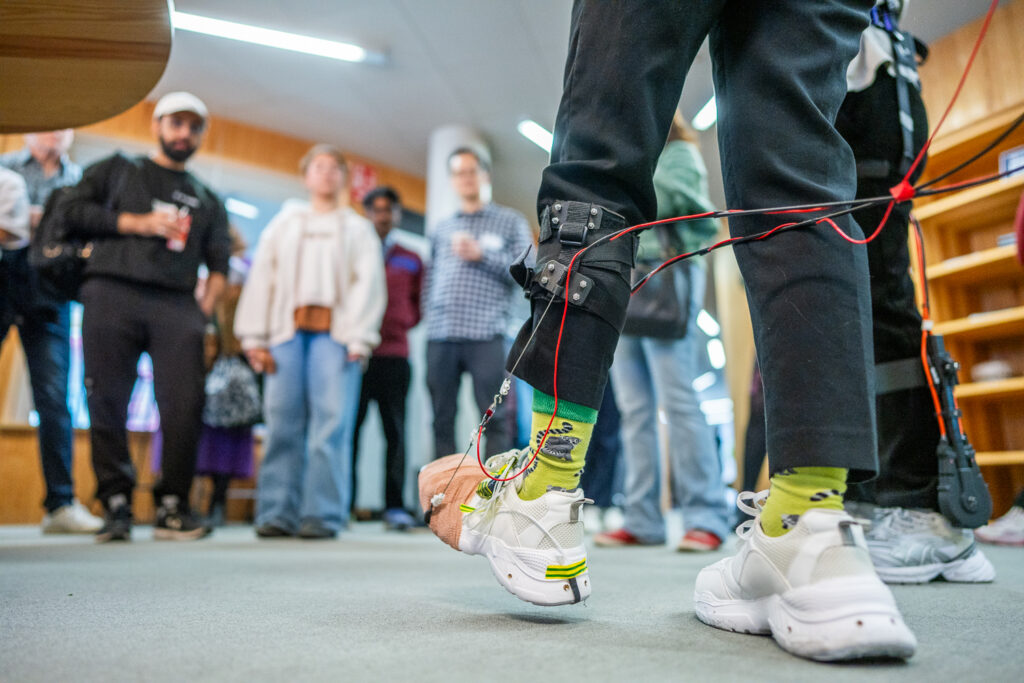 A person wearing white trainers and bright socks has wires attached to their ankles and legs, whilst a group of people in casual clothing watches in the background inside a library or classroom.