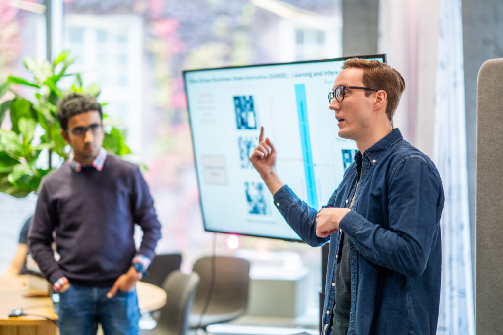 A man in glasses is giving a presentation, gesturing with his hand, whilst another man listens in the background. A large screen displays charts and data in a bright, modern office setting.