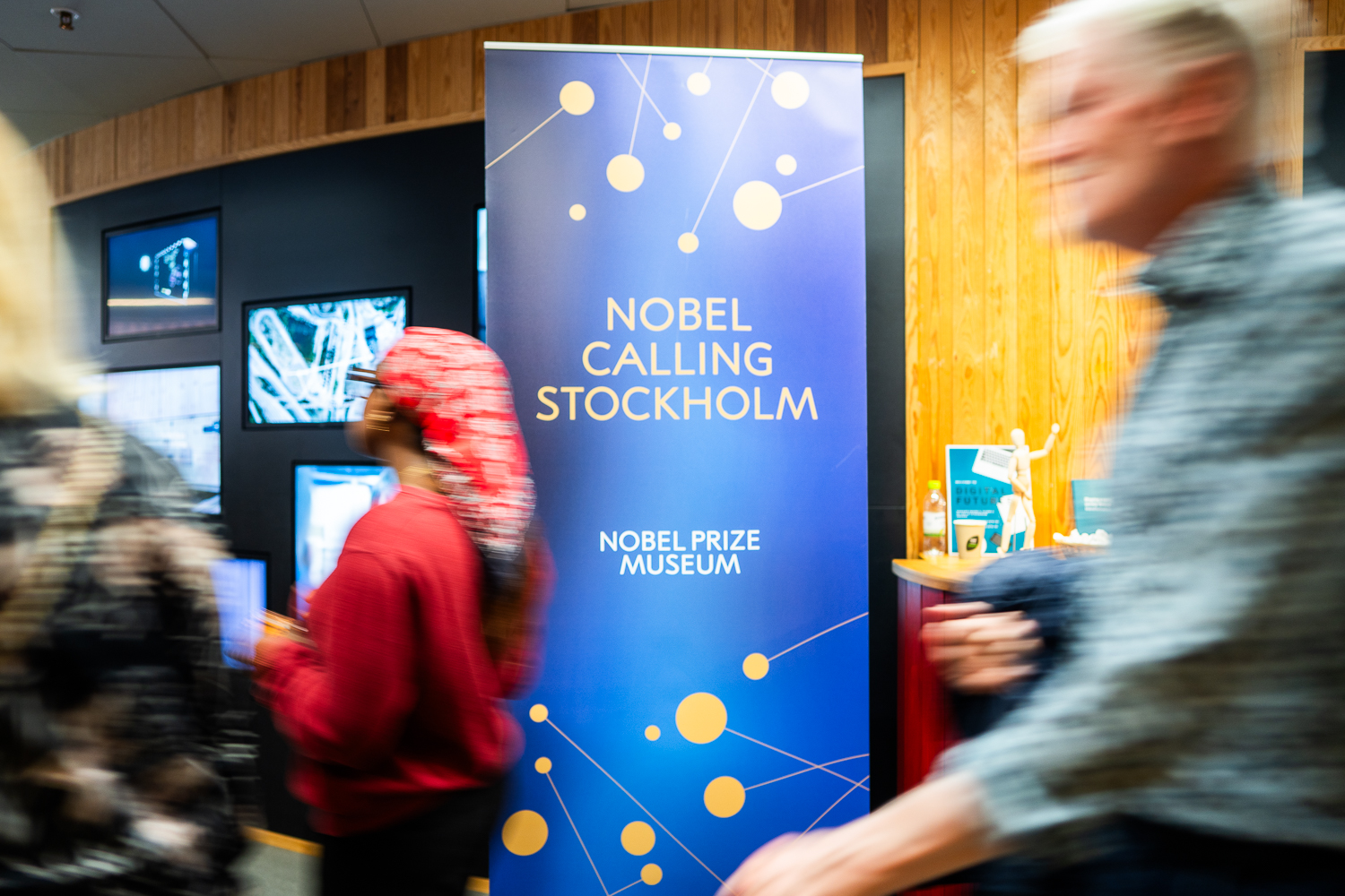 A blue banner reading Nobel Calling Stockholm, Nobel Prize Museum stands indoors, with blurred people walking by and wooden panel walls in the background.