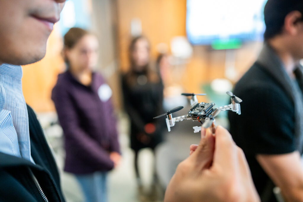 A person holds a small quadcopter drone in their hand, with several people standing blurred in the background indoors.