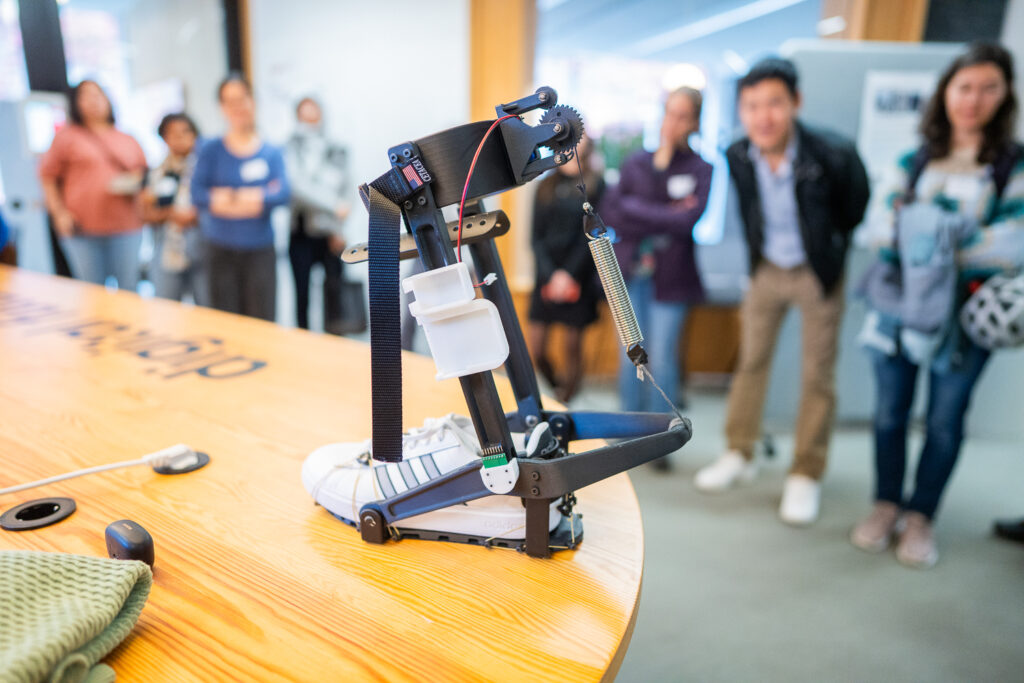 A robotic exoskeleton device is attached to a trainer on a table, while a group of people stands in the background observing the demonstration.