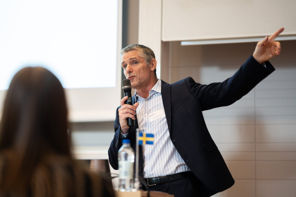 A man in a suit speaks into a microphone while gesturing with his right arm raised, pointing to the side. A blurred person and a small Swedish flag are visible in the foreground.