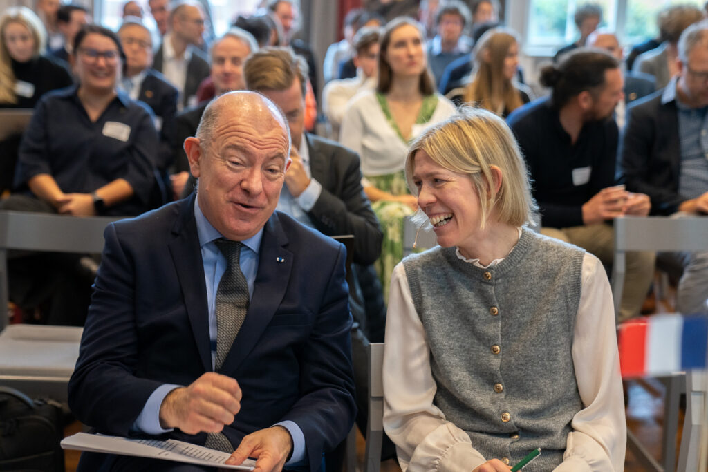 A man in a suit and a woman in a grey waistcoat are sitting and laughing together at an indoor event, with an audience seated behind them, all focused on the front.