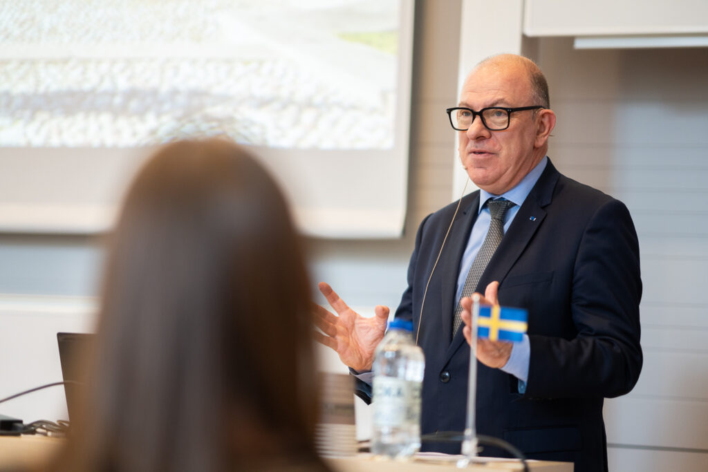 A man in a suit and glasses speaks at a lectern with a small Swedish flag and a water bottle, while a blurred audience member listens in the foreground. A projection screen is visible in the background.
