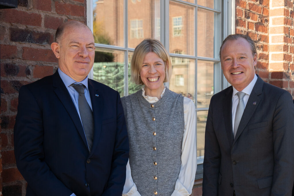 Three people in formal attire stand smiling in front of a brick building with large windows, posing for a group photo outdoors on a sunny day.