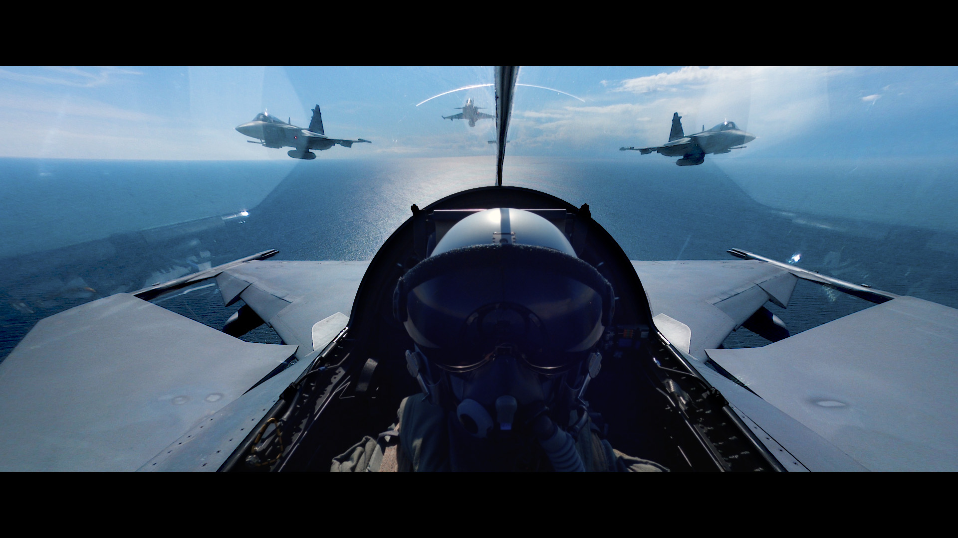 A pilot wearing a helmet and oxygen mask sits in the cockpit of a fighter jet flying over the ocean, with three other jets visible ahead and to the sides in a clear blue sky.