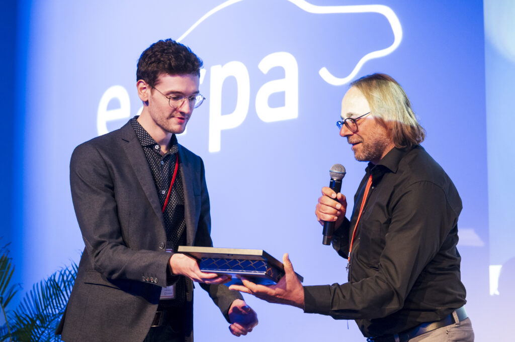 Two men stand on stage; one hands an award in a box to the other, who speaks into a microphone. A blue background with a partial white logo and the word “europa” is visible behind them.