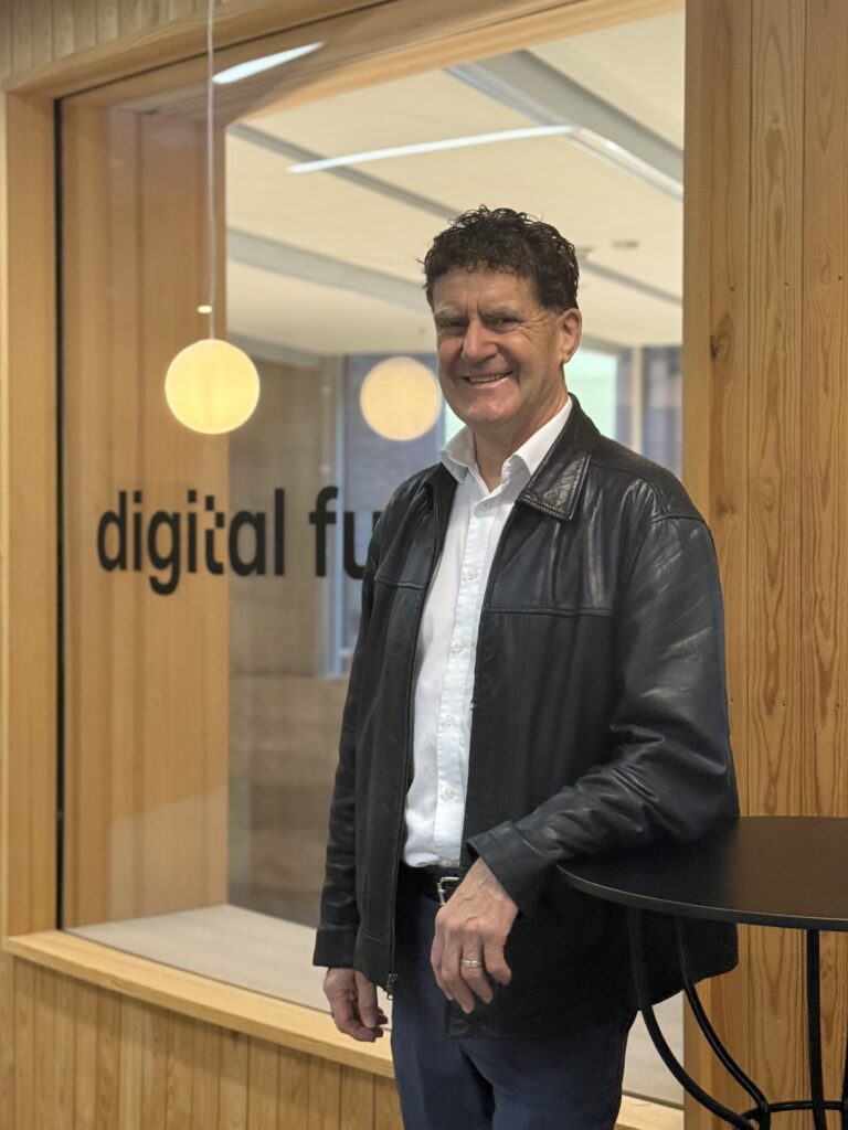 A man with curly dark hair, wearing a black leather jacket over a white shirt, stands smiling next to a round black table in front of a glass wall that reads digital fu.