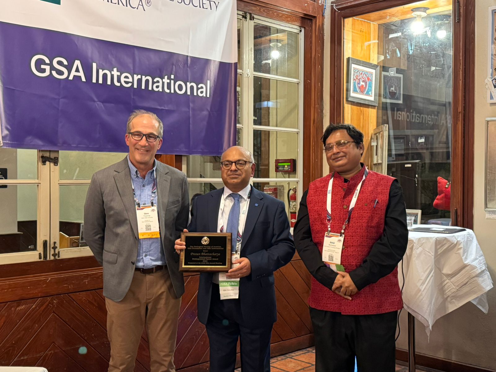 Three men stand indoors beneath a GSA International banner. The man in the centre holds a plaque, and all three are smiling and wearing conference name badges. A glass display case is visible behind them.