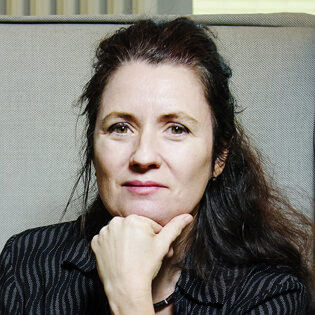 A woman with long dark hair, wearing a black patterned blouse and a watch, rests her chin on her hand and looks at the camera with a neutral expression.