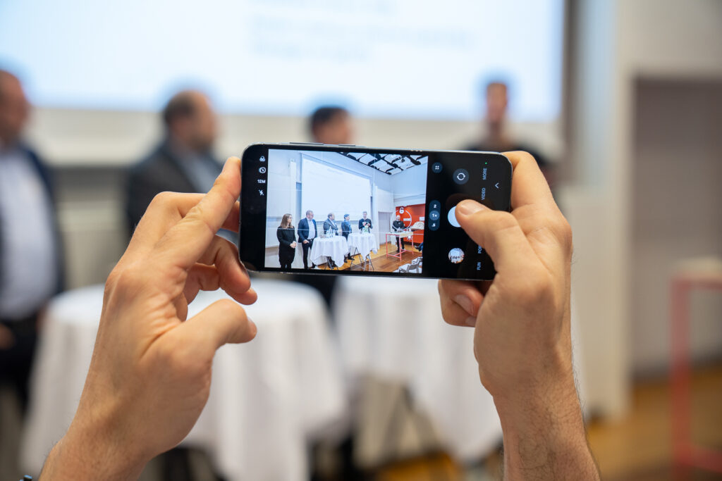 A person holds a mobile phone, taking a photo of four people standing at a table in a conference or event setting. The phone screen clearly shows the group, whilst the background is blurred.