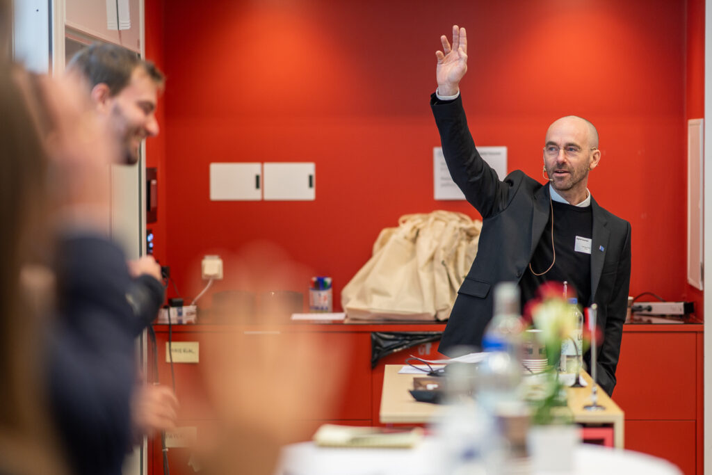 A man in a suit stands and raises his hand, smiling, in a modern room with red walls. Several people in the foreground appear to be engaged, some with raised hands. Office supplies and bags are visible in the background.