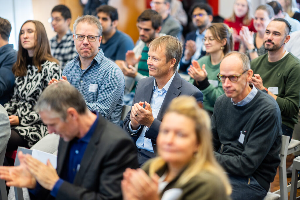 A group of people sits closely together, attentively listening and clapping at an indoor event or conference. Most are wearing name badges and casual to smart-casual clothing, with blurred figures in the foreground.