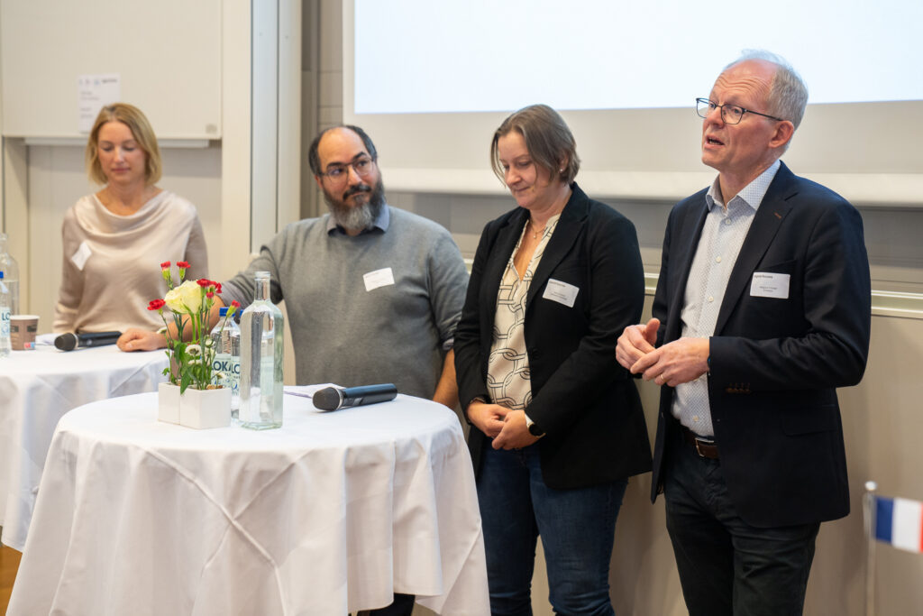 Four people stand at the front of a room near round tables with microphones, water bottles, and flowers, participating in what appears to be a panel discussion or presentation.