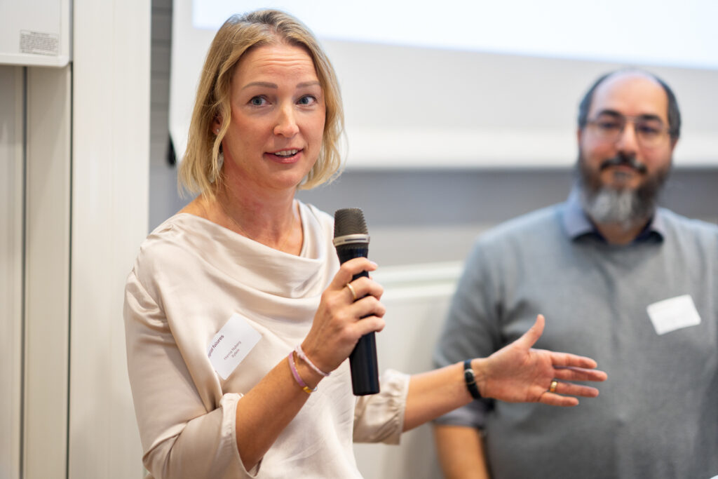 A woman holding a microphone speaks whilst gesturing with her hand; a man with a beard and glasses stands in the background, both wearing name badges in a conference setting.
