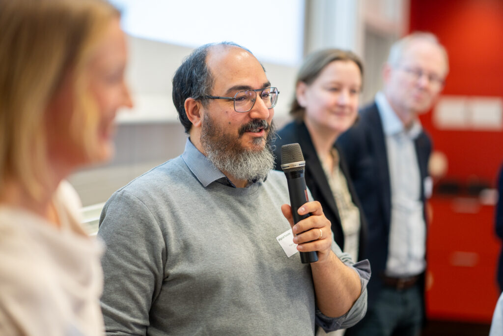A man with glasses and a beard speaks into a microphone whilst standing in front of three other people, all indoors in a brightly lit setting.