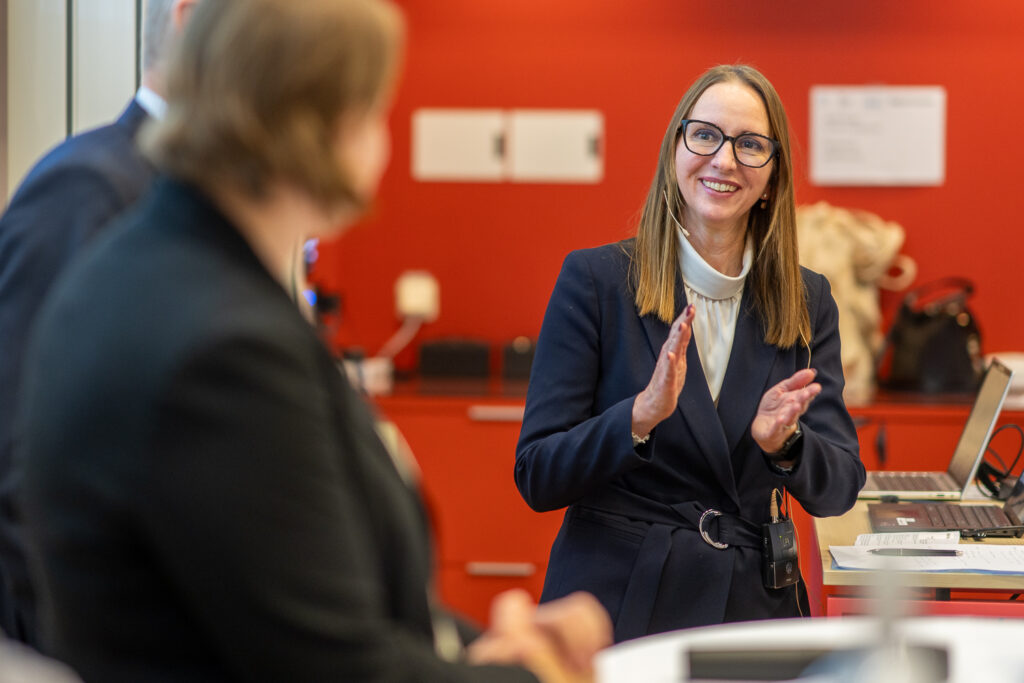 A woman in business attire smiles and gestures with her hands while speaking to colleagues in an office with a red wall in the background. A laptop and bags are visible on the table behind her.