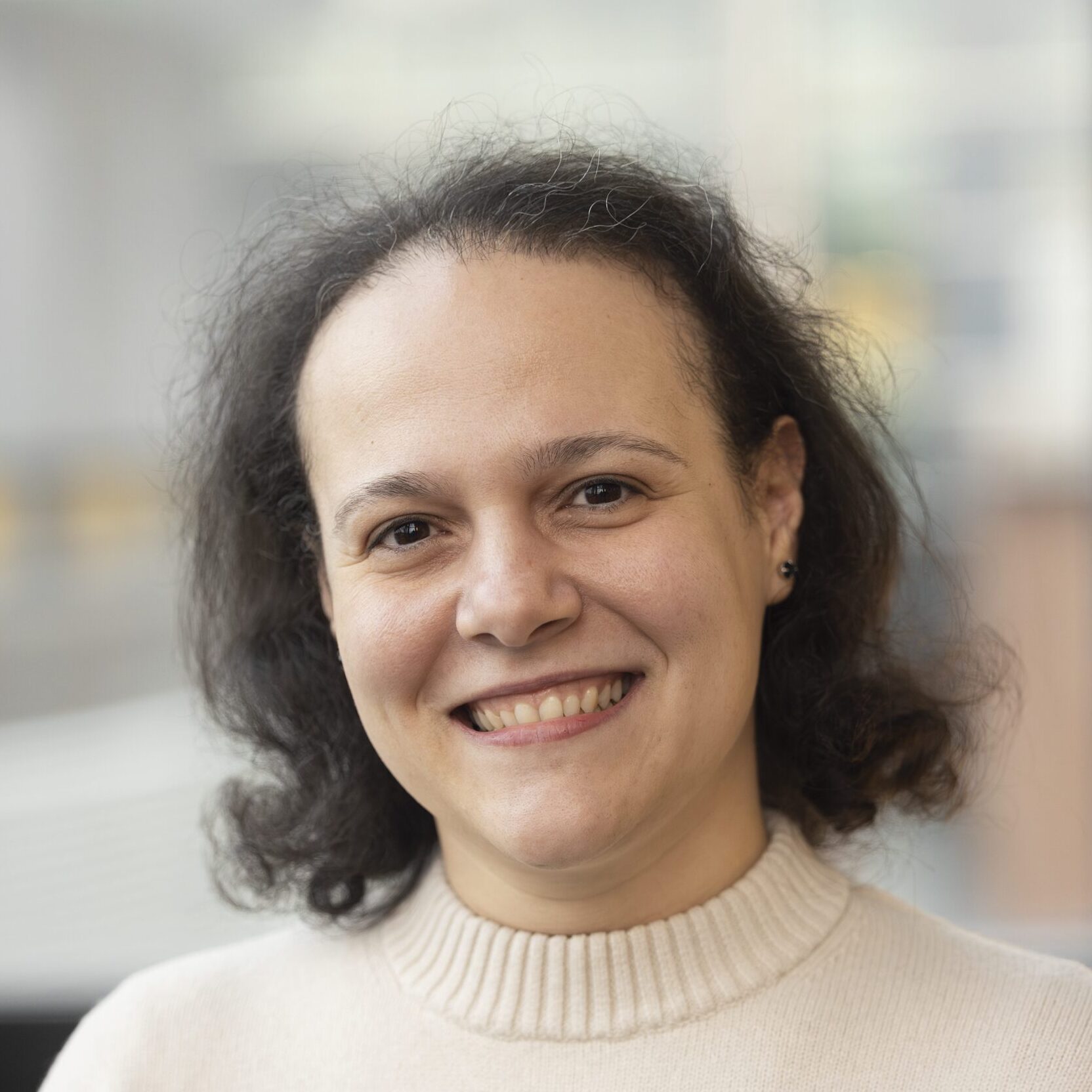 A person with medium-length dark hair smiles at the camera. They are wearing a cream-coloured jumper with black horizontal stripes. The background is softly blurred, suggesting an indoor or urban setting.