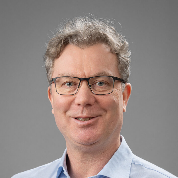 A middle-aged man with short, curly grey hair and glasses smiles at the camera. He is wearing a light blue collared shirt and is posed in front of a plain grey background.