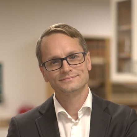 A man wearing glasses and a suit jacket over a white shirt stands indoors, smiling slightly. Bookcases and cupboards are visible in the softly focused background.