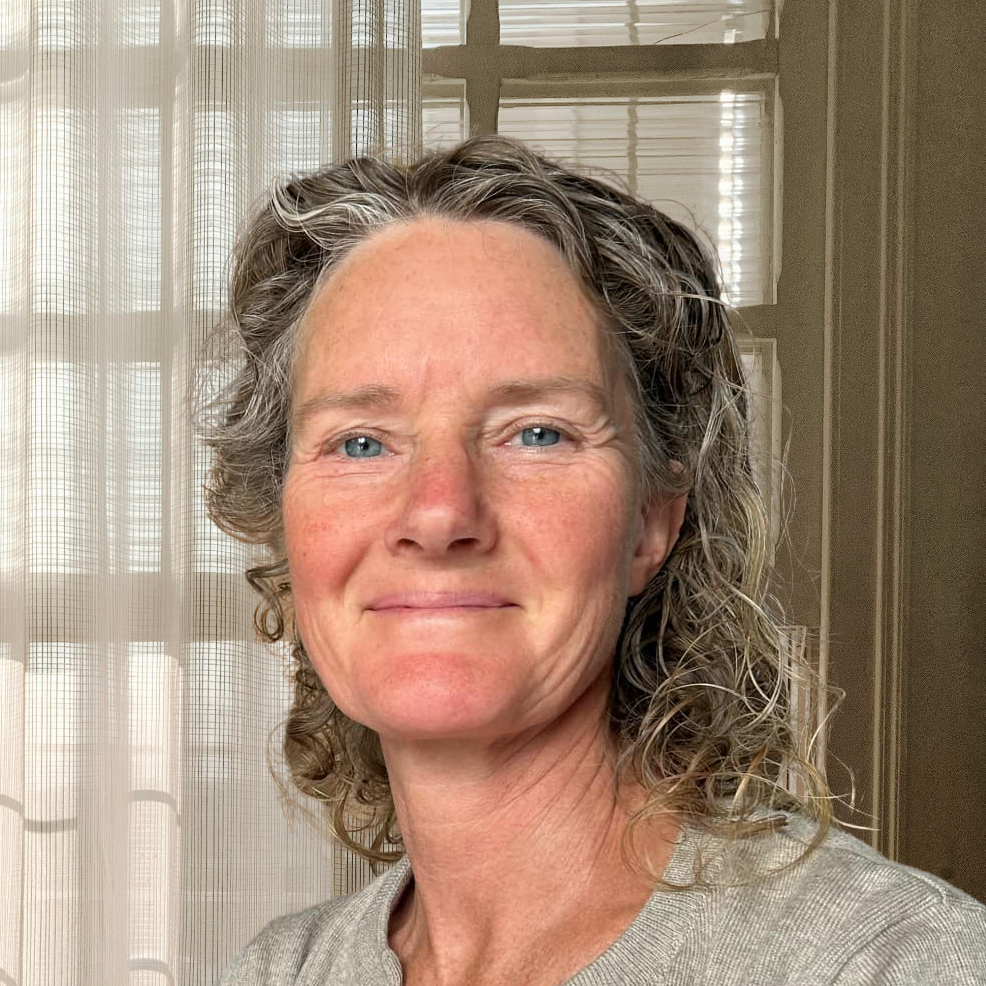 A woman with curly grey hair sits indoors, wearing a grey cardigan over a floral dress. She is smiling gently and holding a mobile, with sheer curtains and window panes behind her. Natural light fills the room.