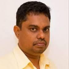 A man with short black hair and a trimmed beard, wearing a light yellow collared shirt, standing against a plain white background, looking slightly to the side.