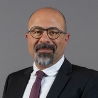 A man with a bald head, glasses, and a short grey beard, wearing a dark suit, white shirt, and maroon tie, poses against a plain grey background.