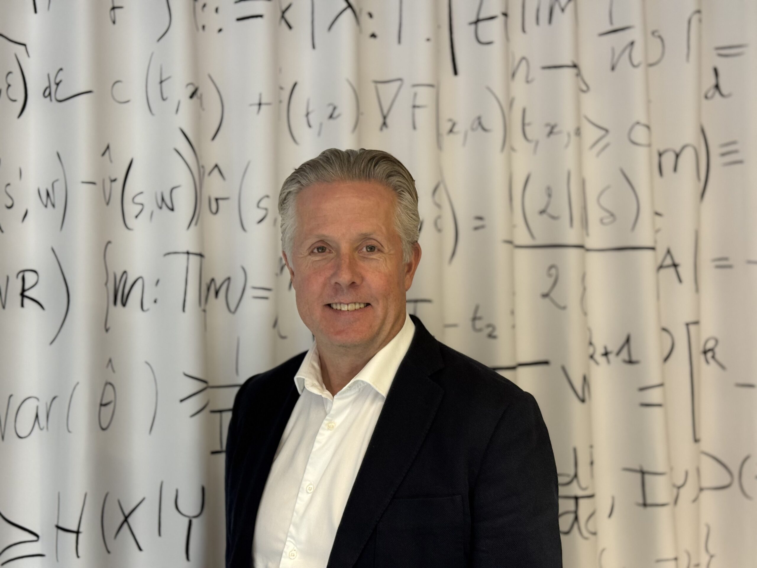 A man in a suit stands smiling in front of a white curtain covered with black mathematical equations and symbols.