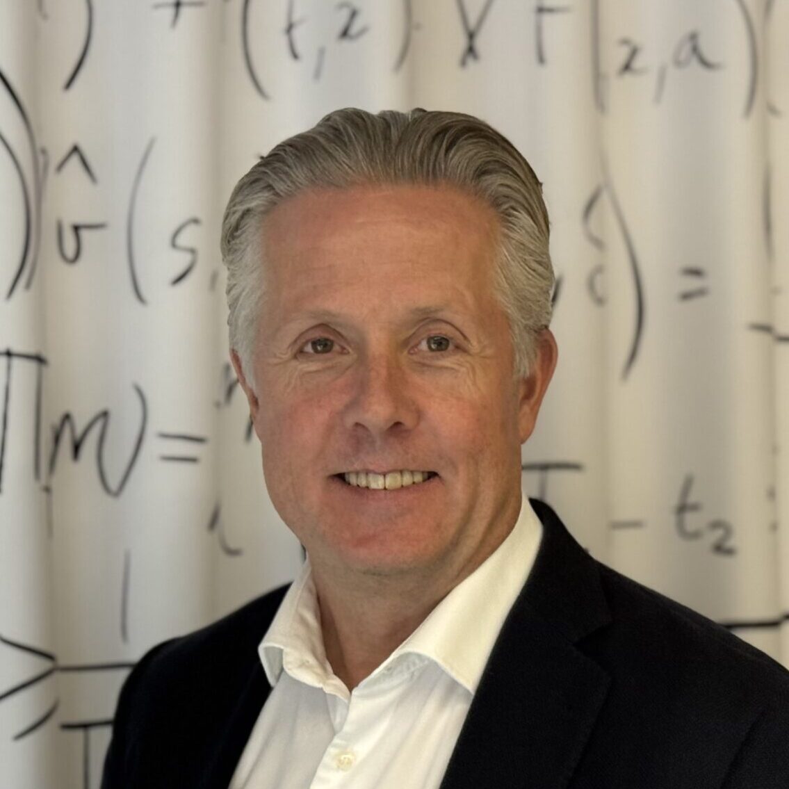 A man in a white shirt and dark blazer stands smiling in front of a whiteboard or curtain covered with mathematical equations and symbols.