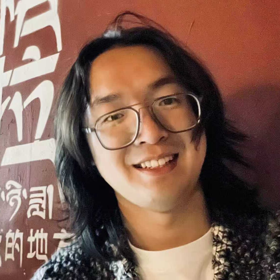 A person with long dark hair and glasses smiles at the camera, wearing a patterned jumper over a white shirt, standing in front of a red wall with large white Chinese characters.