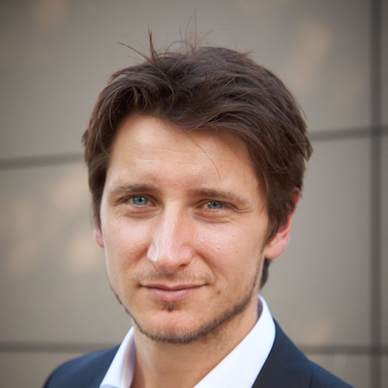 A man with short brown hair and blue eyes wearing a dark suit jacket and white shirt, standing in front of a blurred, neutral-coloured background.