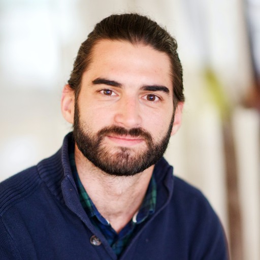 A man with dark hair and a beard, wearing a navy blue jumper over a green checked shirt, is smiling slightly while looking at the camera. The background is softly blurred.