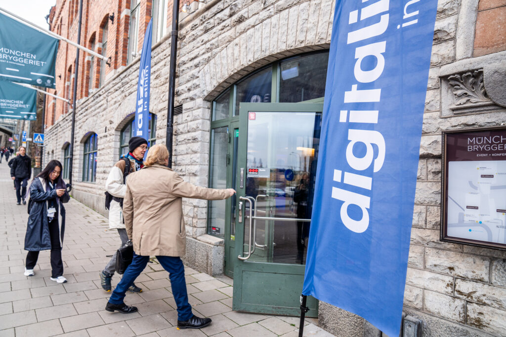 People walk along a pavement next to a stone building with large windows and blue “digitalise” flags. One person is opening a glass door to enter the building. It looks like a business or event venue.
