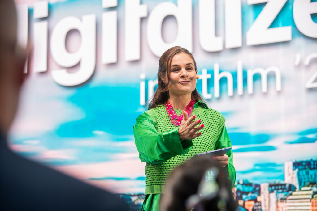 A woman in a bright green outfit and pink necklace speaks on stage, gesturing with one hand and holding notes in the other, with a colourful Digitalise in Sthlm event backdrop behind her.