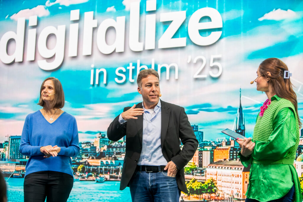 Three people stand on stage in front of a colourful backdrop that reads digitalise in sthlm 25. One man speaks while two women, one with a tablet, listen. A cityscape is visible in the background image.