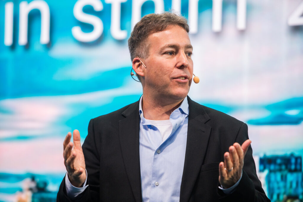 A man in a suit and open-collared shirt speaks on stage, wearing a headset microphone, with his hands raised. A blue and white blurred background with large text is visible behind him.