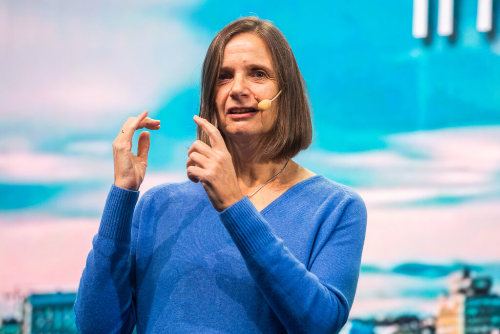 A woman with shoulder-length brown hair, wearing a blue jumper and a headset microphone, gestures with her hands whilst speaking on stage against a blue sky background.