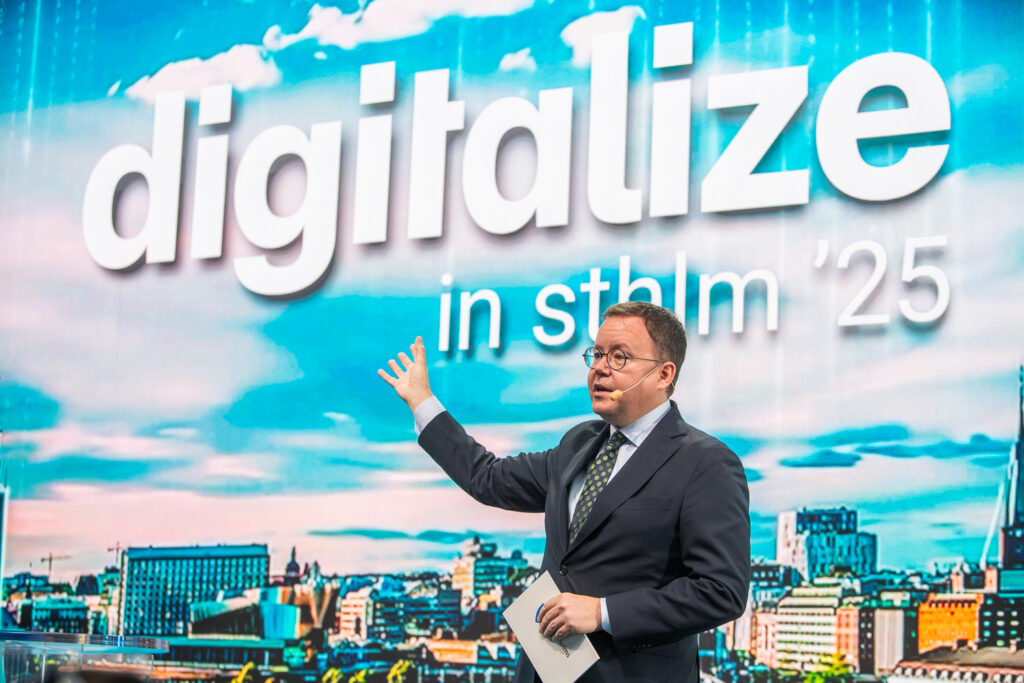 A man in a suit and glasses speaks on stage, gesturing with one hand. Behind him is a vibrant screen displaying the words “digitalise in sthlm ’25” against a cityscape background.