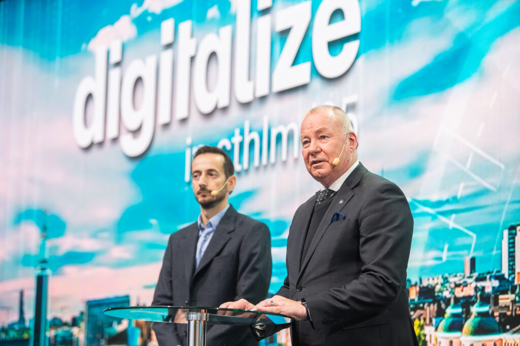 Two men in suits stand on stage with microphones. The background displays the word digitalise in large letters, with a blurred cityscape underneath and a blue sky with clouds.
