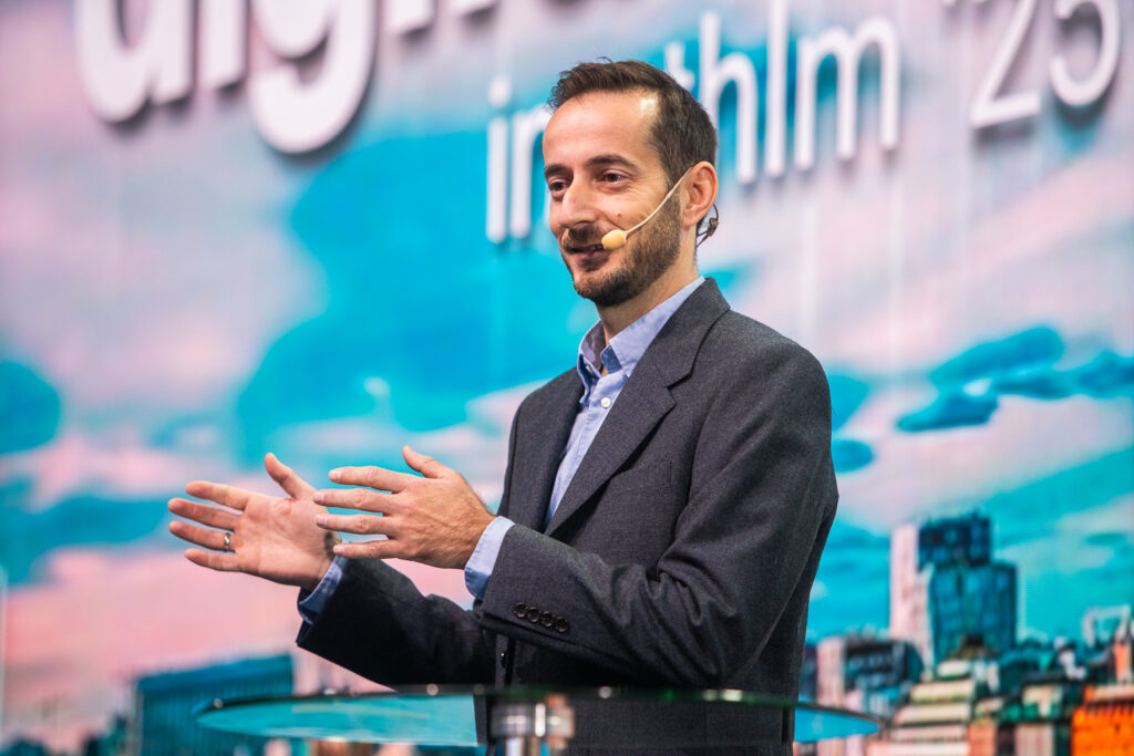 A man in a suit speaks confidently on stage, wearing a headset microphone. He gestures with his hands, standing in front of a blue and pink backdrop with blurred text and cityscape elements.