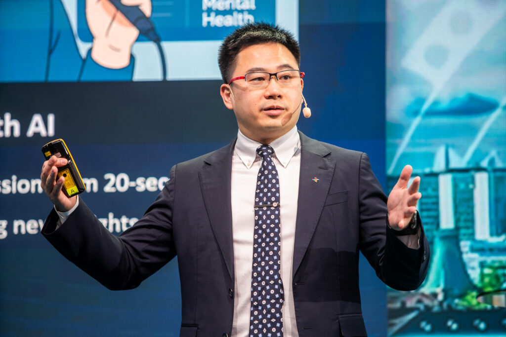 A man in a suit, wearing glasses and a headset microphone, gestures while speaking on stage. He holds a mobile in one hand, with a presentation slide about mental health and AI visible in the background.