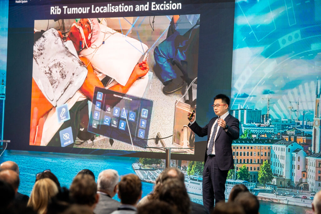 A man in a suit gives a presentation to an audience, standing in front of a large screen displaying a medical image labelled Rib Tumour Localisation and Excision. The audience watches attentively.