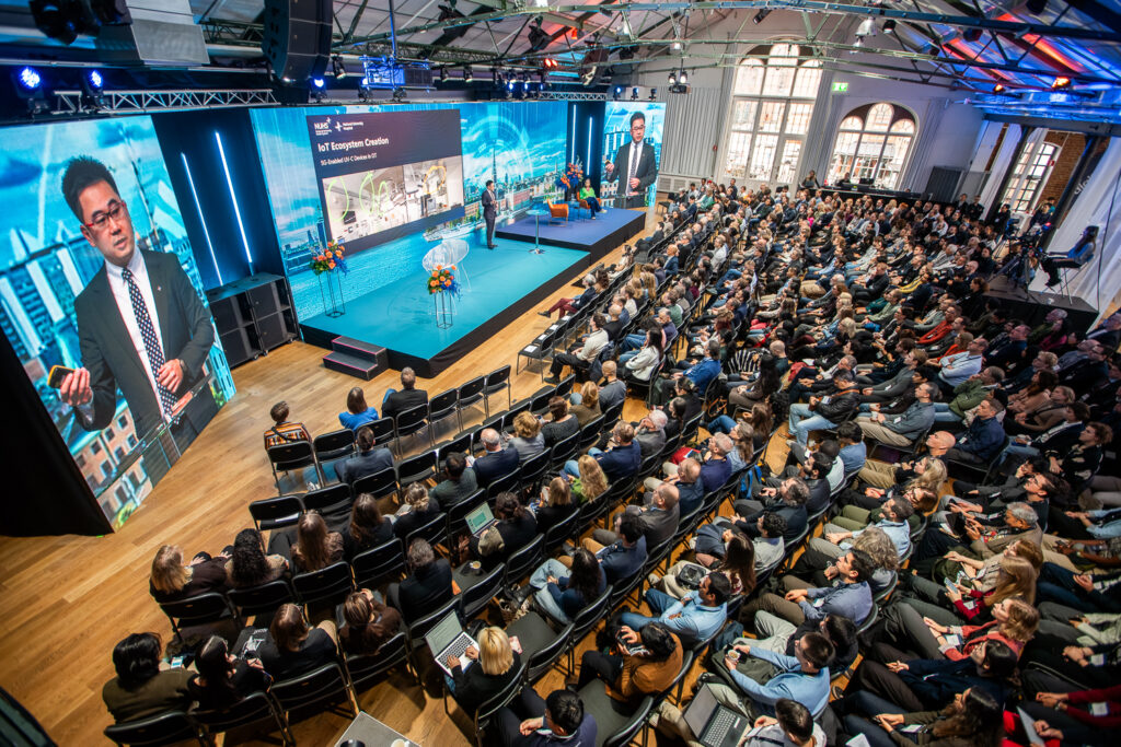 A large audience sits in rows facing a stage where a speaker presents at a conference. A screen behind him displays slides, and two large screens show his image. The venue has high ceilings and bright lighting.