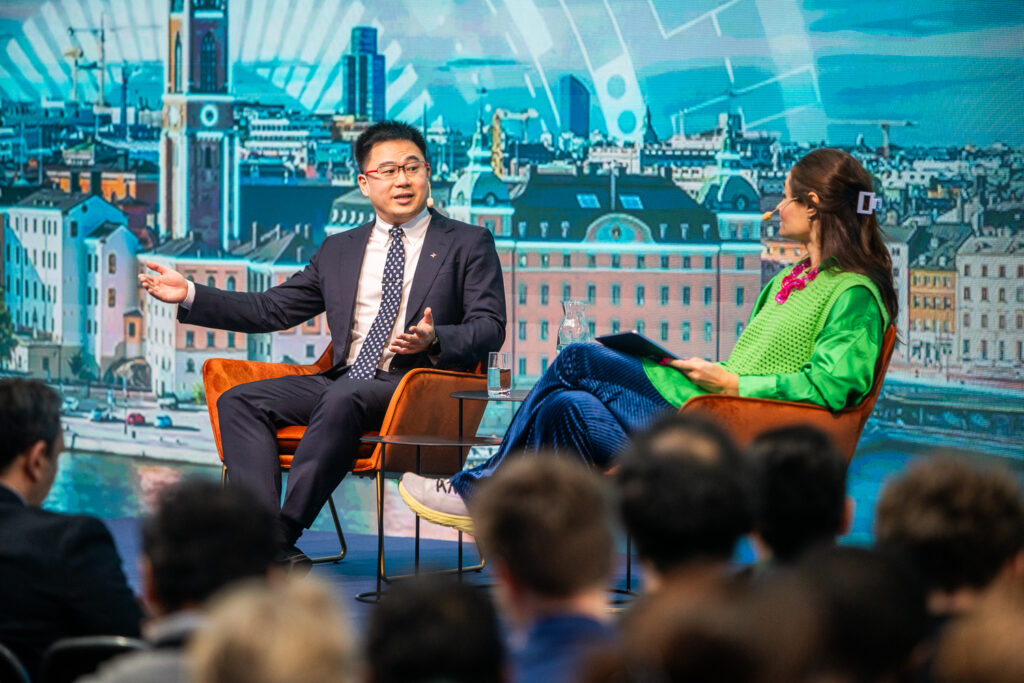 A man in a suit and tie gestures while speaking to a woman in a bright green and blue outfit onstage, in front of an audience. A cityscape backdrop with tall buildings is visible behind them.