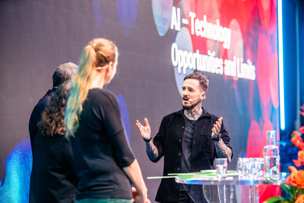 A man speaks animatedly on stage with two other people during an event. The screen behind them displays the text AI – Technology: Opportunities and Limits. Bright lighting and flowers are visible in the scene.