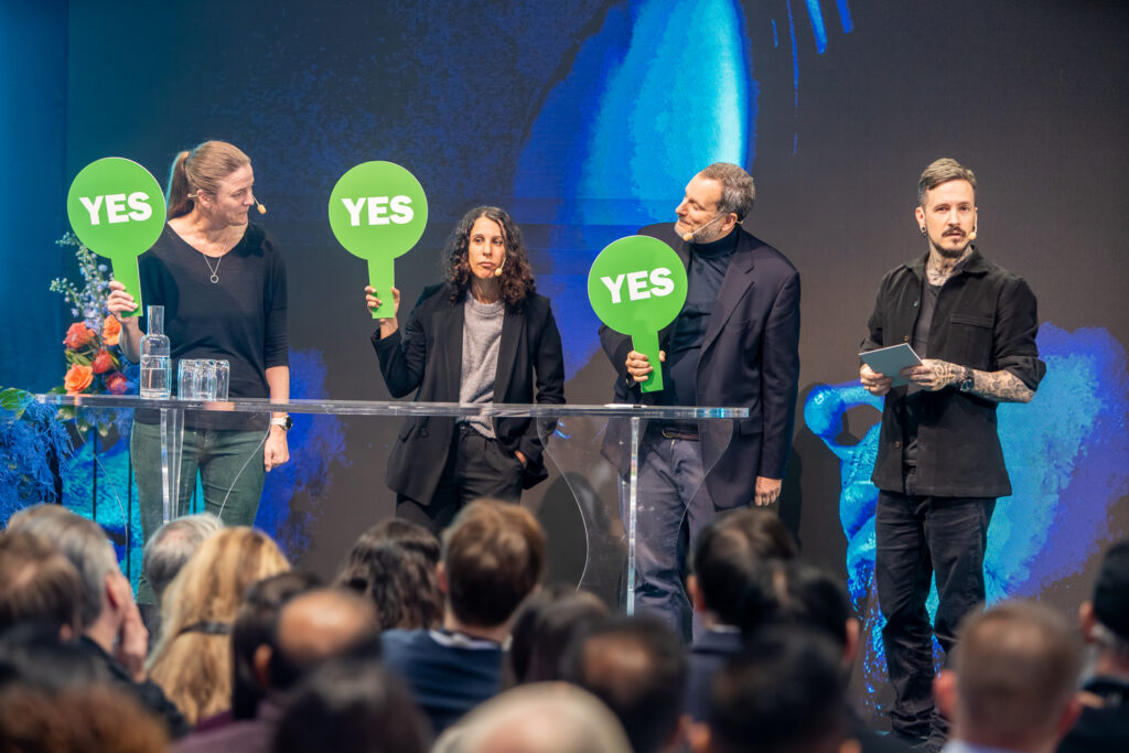 Four people stand on a stage facing an audience, three holding green signs that say YES. One person holds a tablet. Flowers and glasses of water are on a table next to them. A large blue screen forms the backdrop.