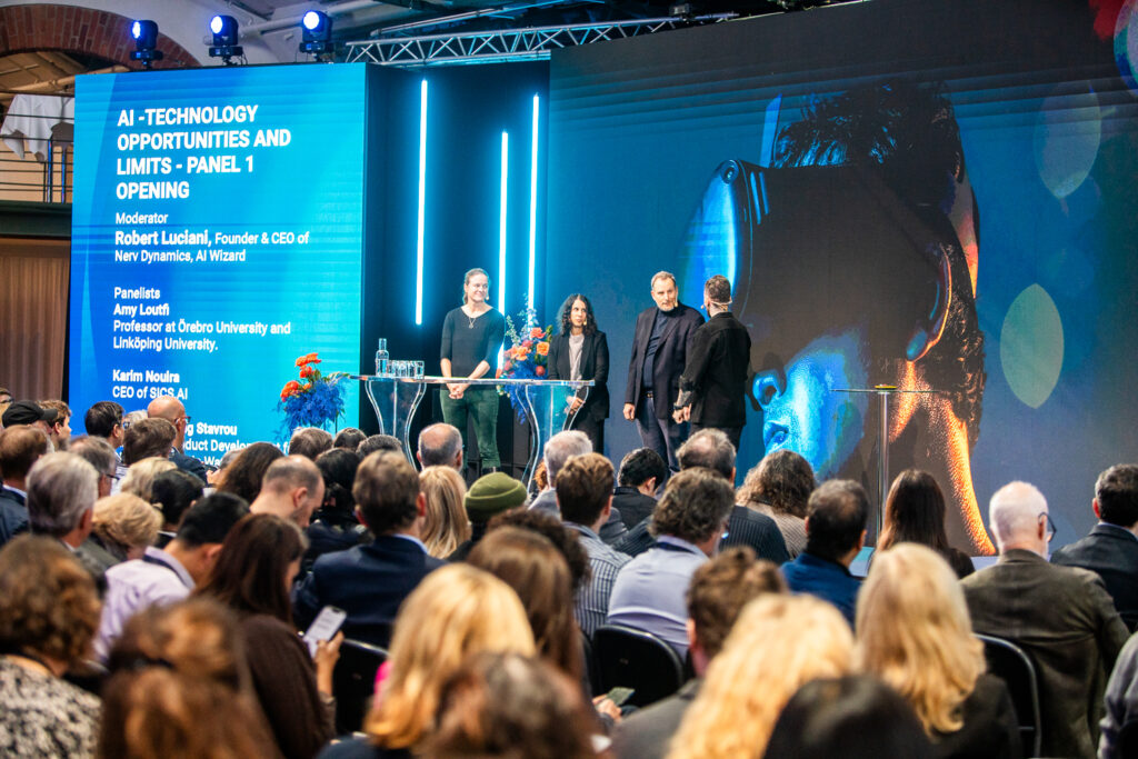 A panel of four speakers stands on stage at an AI technology conference, addressing an audience. Behind them, a large screen displays panel information and an image of a person wearing a VR headset.