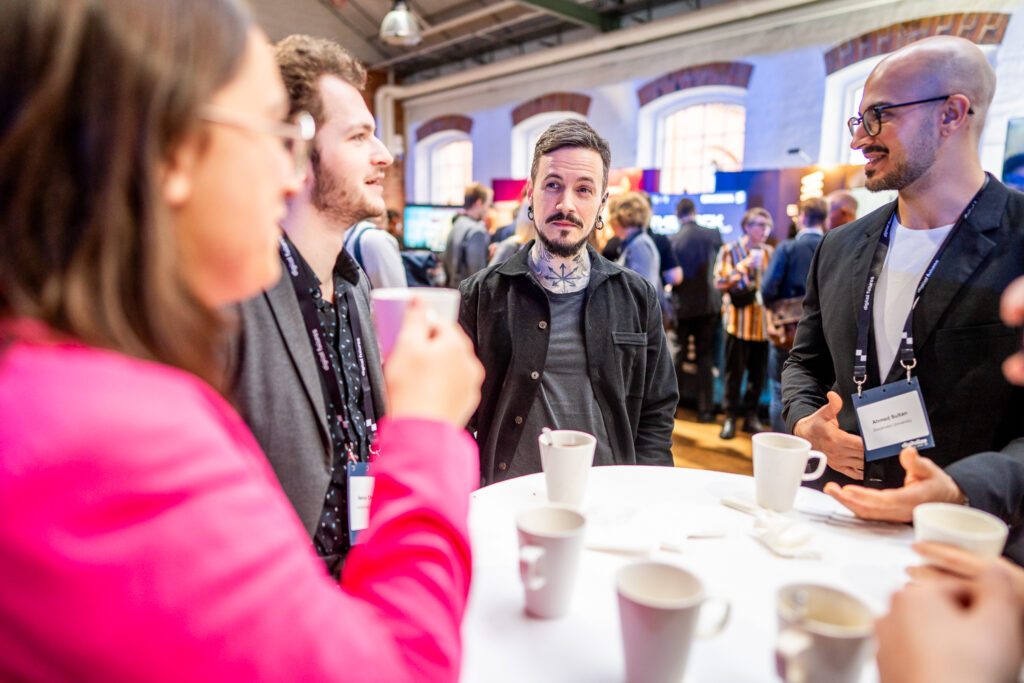 A group of people stand around a table with coffee cups, engaged in conversation at a lively indoor event or conference. Bright lighting and other attendees are visible in the background.