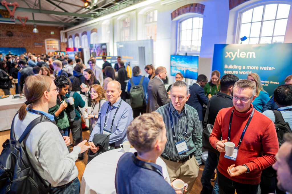 A large group of people gather at a networking event in a bright, spacious venue. Some stand in small groups talking and holding coffee cups, while event stands are visible in the background, including one from Xylem.